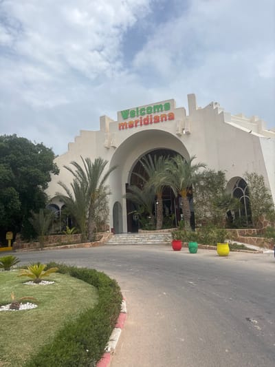 The image shows the entrance to the Meridian hotel in Taguermess, Tunisia. The hotel is a large, white building with an arched entryway. Above the entrance, the words "Welcome Meridiana" are visible in green and red lettering. Palm trees are planted in front of the hotel, and there are several potted plants in colorful containers placed along the driveway. The sky is cloudy.