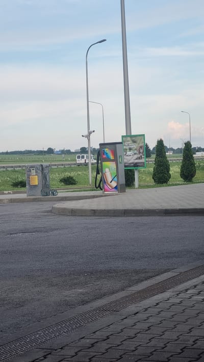 An outdoor scene captures a paved area with a drainage grate running along the edge of a sidewalk. On the sidewalk, two distinct machines are present. To the left, a grey, rectangular machine is covered with a plastic wrap and has a coiled black hose attached. To its right, a taller, grey machine features a colorful advertisement displaying images of car interior cleaning. Beside this machine, a green billboard showcases a hand placing coins into a stack of money next to a car. In the background, a road is visible, with a grassy median and trees lining the roadside. Several white vans are parked on the road. The sky above is mostly clear with some scattered clouds. The location appears to be in Валожынскі сельскі Савет, Belarus.