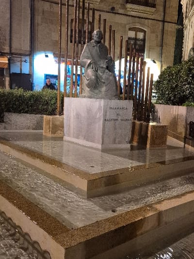 The image depicts a nighttime scene in the Plaza de Fray Luis de León, Salamanca, Spain. Centrally positioned is a bronze statue of a seated male figure, robed and holding a book in his left hand, resting on a rectangular, light-grey stone plinth. The plinth's front face is inscribed with "SALAMANCA AL MAESTRO SALINAS" in capital letters. The statue is framed by an arrangement of numerous slender, dark brown vertical poles.

In the immediate foreground, a multi-tiered fountain is visible, constructed from light-toned concrete or stone with distinct speckled brown granular edges on each tier. Water flows gently across these flat surfaces, creating ripples. Behind the statue, dense green bushes form a low hedge. Beyond the foliage and poles, a traditional stone building with a single window is visible to the right. To the left, an illuminated shopfront, possibly a bar or restaurant, stands out, with a faint blue glow emanating from its interior. A lone male figure in a dark jacket is partially visible in the background, positioned to the left of the statue, standing behind the bushes and poles near the illuminated storefront. The scene is lit by artificial urban lighting.