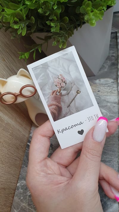 In this close-up shot, a hand with polished pink and white nails holds a rectangular white card. The card features a photograph on its left side, depicting a person's arm in a pink knitted sweater holding a small bouquet of pale pink roses and a miniature spoon. The background of the photo is a soft, draped white fabric, suggesting a cozy or intimate setting. To the right of the photograph, the card displays text in black, vertical lettering. The visible text reads "Красота - это ты" which translates to "Beauty is you" in Russian, followed by a small black heart icon.

The hand holding the card is positioned in the foreground, with its fingers gently gripping the card's edges. In the background, elements of a domestic setting are visible. To the left, a leafy green plant adds a touch of nature. Behind the plant, a light grey, angled surface suggests a box or piece of furniture. The surface beneath the card and other objects appears to be a wooden table. A small, cream-colored object with rose gold spectacles is partially visible in the lower foreground, adding a whimsical touch. The lighting suggests an indoor setting, possibly during the daytime, with soft illumination. The overall impression is one of gentle aesthetics and personal sentiment, possibly related to beauty or self-appreciation, given the text on the card. The location context of Aprelevka, Russia, does not add specific visual cues to the scene itself.