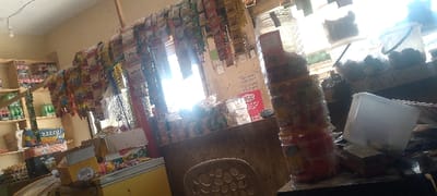 The image displays the interior of a small retail shop situated in Danbare, Nigeria. The scene is illuminated by strong natural light originating from an exterior opening on the right side, resulting in significant overexposure in that area. An exposed incandescent light bulb is suspended from the ceiling in the upper-left quadrant.

The shop's walls are light-colored. Along the left wall, multi-tiered wooden shelves are stocked with bottled beverages, identifiable by red and green labels. Below these shelves, various packaged goods and cardboard boxes, one featuring "ZIZZOU" branding, are arranged. A yellow chest freezer or cooler is partially visible in the lower-left foreground.

A counter or display surface spans the mid-ground. Above this counter, multiple horizontal lines suspend numerous small, individually packaged snack items. Behind the counter, additional packaged goods are stacked, and a white plastic chair with a perforated back is positioned.

On the right portion of the counter, a stack of clear plastic containers is present. Behind these, several large clear plastic buckets with handles hold dark, indistinct contents. A smaller, opaque white plastic container rests on the counter. The wall behind the right counter exhibits faint handwritten text and a red poster or sign. No individuals are present within the depicted scene.