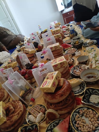 The image depicts a table laden with food and drinks, likely for a gathering or celebration. There are various types of bread, including large, circular loaves with intricate patterns, and smaller, crescent-shaped pastries. Several plates contain nuts, some appearing to be pistachios. There are also bowls of fruit, including grapes and peaches, and slices of watermelon. Packaged snacks, possibly cookies or biscuits, are stacked on the table, some with prominent branding. Bottles of liquid, possibly juice or water, are also present. Decorative elements like small golden teapots and translucent gift bags with pink ribbons are visible. The table is covered with a patterned cloth. The background suggests an indoor setting, with hints of furniture and a wall. The scene appears to be from a city in Kyrgyzstan, specifically Osh. The overall impression is one of abundance and hospitality.