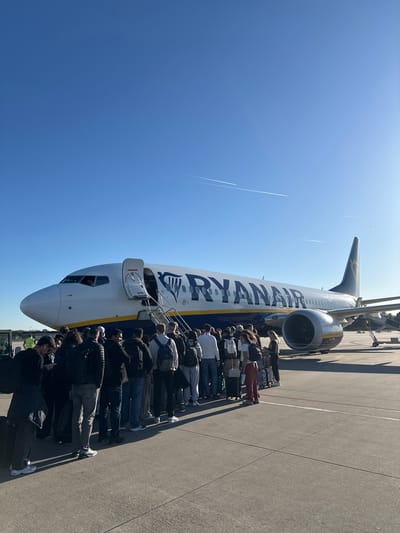 This sunlit image captures a long queue of passengers boarding a Ryanair aircraft on an airport tarmac in Weeze, Germany. The perspective is taken from slightly behind and to the side of the queue, looking towards the front of the airplane.
The main subjects are a large group of people and a white and blue Ryanair airplane, likely a Boeing 737, with "RYANAIR" clearly visible along its fuselage and the "RYR" logo on the tail. The passengers, dressed in casual attire and carrying various bags and luggage, are lined up in single file, extending from the front door of the aircraft where a set of mobile stairs is connected. They appear to be waiting to ascend the stairs and enter the plane.
The scene is an airport apron under a bright, clear blue sky. Strong sunlight casts prominent shadows of both the aircraft and the people onto the asphalt tarmac, indicating a sunny day, likely during midday or early afternoon. Several thin, white contrails are visible high in the sky. The ground shows typical airport markings. No strong emotions are discernible from the passengers, who seem to be engaged in the routine process of boarding.
