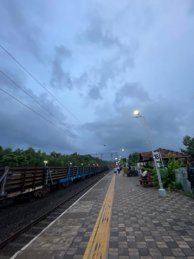 The image depicts a train station platform in Sircaim, India, under a cloudy sky, suggesting it might be late afternoon or evening. The platform is paved with dark and light grey square tiles, with a distinct yellow tactile paving strip along the edge. To the right of the platform, a train with blue carriages is visible on the tracks. Overhead, power lines crisscross the sky.

On the platform, several people are visible, some sitting on benches near a building with a tiled roof. A lamppost illuminates the scene, casting a warm glow. A sign with the number "18" in blue is mounted on a pole, likely indicating a platform number. The background is filled with lush green trees, adding to the natural setting. The overall mood appears calm, with people seemingly waiting for a train or having just arrived. The weather appears overcast, hinting at a possibility of rain or that the day is drawing to a close.