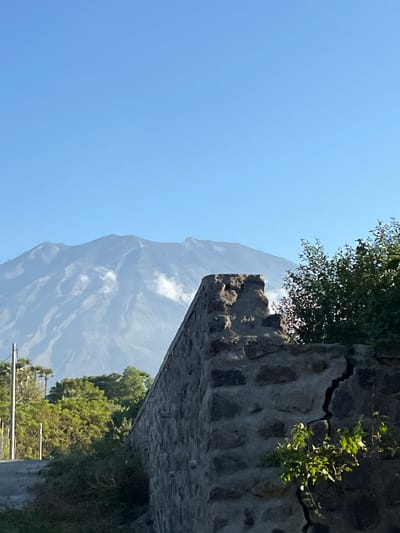 This media file is an image of a mountainous landscape in Kubu, Indonesia. The image is oriented vertically. In the foreground, there is a stone wall with a large crack running through it. Green foliage grows from the crack and from the top of the wall. Beyond the wall, a large, imposing mountain looms, its slopes partially obscured by soft white clouds. The sky is a clear, bright blue, suggesting a sunny day. In the lower right of the image, the tops of trees and what appear to be utility poles are visible, hinting at a road or pathway near the foreground. The scene conveys a sense of natural grandeur and perhaps the ruggedness of the terrain. There is no visible text in the image.
