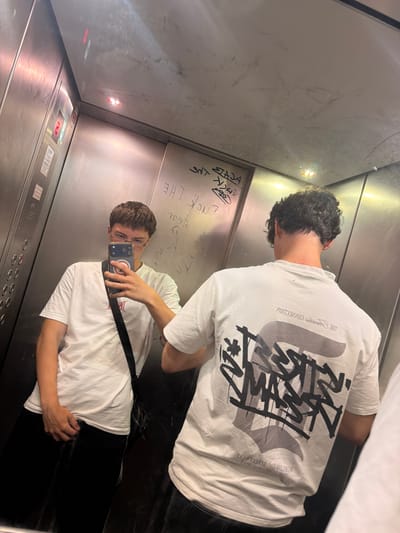 In an elevator with brushed metal walls, a young person wearing glasses, a white t-shirt, and black pants is taking a selfie in a phone's reflection. The elevator has graffiti on the walls and a red illuminated button panel. The person is holding a phone and has a dark crossbody bag. Another person wearing a white t-shirt is partially visible in the reflection, facing away from the camera. The elevator appears to be located in an urban environment, potentially in a building within the city of Dorsten, Germany, suggested by the presence of an elevator and graffiti typical of such settings.