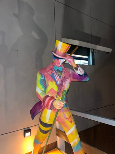 A stylized, multi-color statue of a male figure is prominently displayed against a flat, light grey wall. The figure is depicted wearing a top hat, a suit jacket, a bow tie, and trousers. The entire ensemble is painted with a mosaic of bright, varied colors and patterns, including stripes, geometric shapes, and abstract designs in pink, yellow, blue, green, and orange. The statue's right hand is positioned at the brim of its vertically striped top hat, while its left hand firmly holds a green cane with a spherical pommel. A defined shadow of the figure is cast on the wall to its left. Below the statue on the left, a square, wall-mounted light fixture emits a warm, orange illumination onto its lower left leg. In the upper right background, a rectangular window or opening is partially visible beneath a horizontal overhang. A wooden railing or barrier extends across the lower portion of the frame, suggesting the statue's placement on an elevated platform or near a stairway. This scene is located in Ayamonte, Spain.