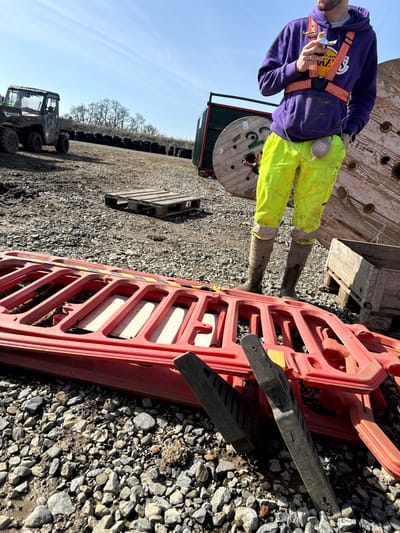 The image depicts an outdoor industrial or construction site on a clear day in Baldurgan Hill, Ireland. The ground consists of coarse gravel and dirt. In the foreground, two red plastic temporary barriers are stacked horizontally, with sections of yellow and black hazard tape visible on the top barrier. Two dark grey plastic barrier feet are angled against them in the lower right.
A person is centrally positioned, visible from the chest down, wearing a purple hooded sweatshirt with a yellow Lakers logo, an orange high-visibility harness, bright neon yellow work pants, and dark brown muddy work boots. The individual holds a clear bottle with an orange label in their right hand. A grey JBL portable speaker is clipped to the person's right hip, connected by a white wire.
In the midground to background, a green utility terrain vehicle (UTV) is parked on the far left. To its right, two wooden pallets are stacked. Further right and in the background, multiple large wooden cable reels are present, with one clearly marked "222 CABLE." A green industrial container or trailer is partially visible behind one of the cable reels. A line of black stacked tires forms a distant barrier. A small wooden crate or pallet box is situated to the right of the person's feet.