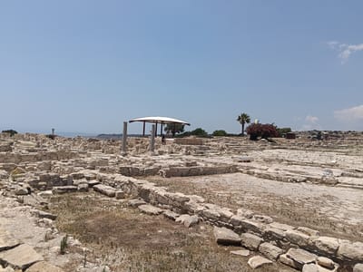 The image depicts an archaeological site in the city of Episkopi Cantonment, Sovereign Base Areas of Akrotiri and Dhekelia. The scene is dominated by ancient stone ruins, with walls and foundations made of rough-hewn blocks scattered across a dry, dusty landscape. In the mid-ground, a white canopy supported by columns offers shade over a section of the ruins, and a single person can be seen standing underneath. A palm tree and a bush with red flowers are visible to the right of the canopy, and another palm tree further to the right. The sky is clear and blue, with a few wispy clouds in the distance. The overall impression is one of a sun-drenched, historical site.