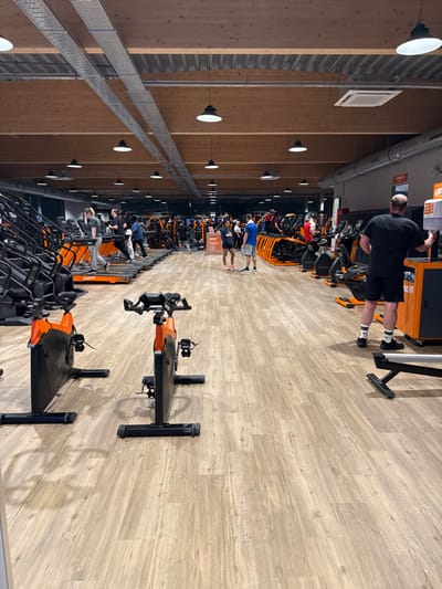 Inside a well-lit gym with a high ceiling and light wood paneling, rows of stationary bikes are arranged. People are working out, with one individual standing on a machine, adjusting it. The gym has overhead lighting fixtures and cable trays running along the ceiling. The floor is a light, wood-patterned surface. The bikes are predominantly black and orange.