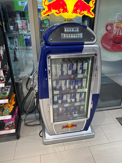 An upright, blue and silver Red Bull branded cooler, designed to resemble a vintage gasoline pump, stands on a light grey tiled floor inside a convenience store. The cooler's top section features a yellow sign with two red charging bulls, the Red Bull logo, and a digital-style display below it reading "NEED A REFUEL?". The main body of the cooler is a glass-fronted refrigeration unit, fully stocked with multiple rows of blue and silver Red Bull energy drink cans. A black hose with a silver nozzle, imitating a fuel pump hose, hangs from the left side of the cooler. To the immediate left, a black metal retail shelving unit is partially visible, holding various packaged snacks, some with Cyrillic text and a product labeled "JUMP." In the background, large glass windows and doors reveal reflections of interior elements and exterior details, including an "OMV" logo, suggesting the location is within an OMV gas station shop. The scene is located in Gorno Aleksandrovo, Bulgaria.