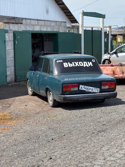 A teal-colored Lada 2107 sedan is parked on a gravel surface in front of a partially open garage with green double doors. The car has a white sticker on its rear window that reads "ВЫХОДИ" (vykhodi), which translates to "Get out" or "Exit" in Russian. The license plate is visible and reads "A 146PB 172 RUS". In the background, a rusty trailer is visible, along with a silver vehicle and a green metal fence. The scene appears to be in an outdoor, possibly industrial or rural, setting. The sky is clear and blue, indicating fair weather. The overall impression is of a parked vehicle in a utilitarian area.