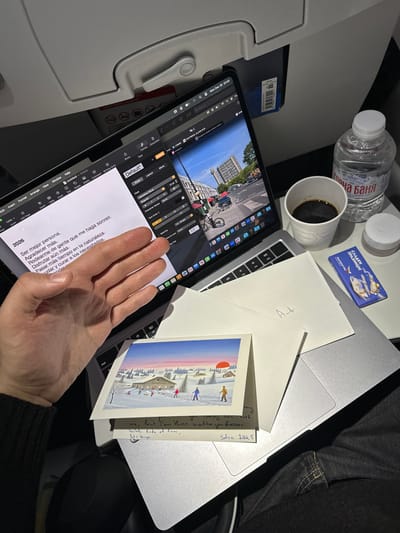 The image captures an in-flight scene with an open laptop displaying photo editing software alongside a tablet with a text document in Spanish outlining goals for 2026. Adjacent is an opened Christmas card with a winter landscape illustration and a handwritten message in English, dated "Satra 2015". Beverages include a bottle of "Горна Баня" water and a cup of coffee. Also present are a chocolate bar branded "СЛАДКИ ПРАЗНИЦИТ" and envelopes. A person's hand rests on the tablet screen, and the scene is illuminated by artificial light.