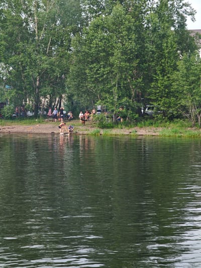 The image depicts a riverside scene in Ермолаевский Затон, Russia. On the grassy bank, several people are gathered under the shade of tall green trees. Some are sitting on chairs or the ground, while others are standing or walking. A few individuals are near the water's edge. In the foreground, the dark, rippling surface of the water fills the frame. The overall atmosphere suggests a casual gathering or outing by the river.