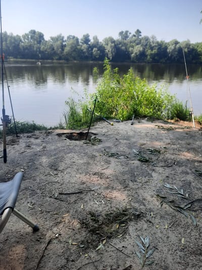 The image depicts a natural scene along the bank of a body of water, likely a river, in Pukhivka, Ukraine. In the foreground, a sandy area with scattered leaves and branches is visible. A portion of a grey folding chair is present on the left side of the frame. Several fishing rods are planted in the ground near the water's edge, suggesting a fishing activity. One fishing rod on the left is closer and shows a fishing reel. A notable feature in the center of the foreground is a dark, irregularly shaped hole in the ground, possibly a burrow or a dug-out area. Further back, lush green vegetation grows along the bank, with tall grass and bushes. The water is calm and reflects the trees lining the opposite bank, creating a dense green treeline against a clear blue sky. A small boat with one person is visible in the distance on the water. The overall atmosphere is peaceful and indicative of an outdoor recreational setting.