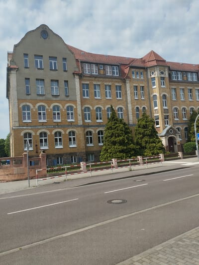 The image depicts a large, ornate, multi-story building, likely a school or administrative building, in Bitterfeld-Wolfen, Germany. The building has a mixed architectural style with a tan-colored brick facade, arched windows on some floors, and a contrasting grey section on the left side with rectangular windows. The roof is red tile. Two large, conical evergreen trees are positioned in front of the building, partially obscuring the main entrance.
A low, red brick wall with a white and red striped railing runs along the front of the property. A paved road with white lane markings is visible in the foreground. There is no visible human activity. The sky is partly cloudy.