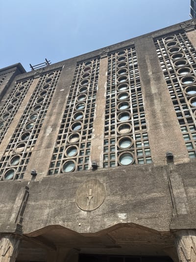 The image shows a low-angle shot of a building facade in the Hongkou District of China. The building is constructed of gray concrete and features a repetitive pattern of circular windows set within a grid of rectangular openings. The facade is stained and weathered, suggesting age and exposure to the elements. On the right side of the building, there is a circular plaque with an emblem of some kind; there is a dark doorway nearby. The sky is visible in the upper left corner of the frame and is a pale blue. The lighting is diffused. A portion of the building's upper corner is also visible.