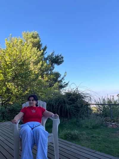 The image presents an outdoor scene located in the Merom HaGalil Regional Council, Israel. An adult female is seated in a white, slatted Adirondack-style chair positioned on a wooden deck. She is wearing a red short-sleeved t-shirt with a blue and white star-shaped emblem on the chest, and light blue and white vertically striped pants. Her dark hair is styled around her shoulders. She has small hoop earrings and a ring on her left ring finger. Her right hand, resting on the chair's armrest, holds a small, clear object, possibly a glass. She faces slightly to the right of the viewer, with her head subtly elevated.
The background features lush green vegetation, including tall, sun-drenched trees with various shades of green foliage on the left and center-left. Dense, darker green bushes, some with visible thorny branches, extend across the mid-ground. A dark metal railing is present in the far right mid-ground, suggesting a perimeter or overlook. Beyond the vegetation, a lighter, hazy horizon is visible under a clear, unblemished blue sky that occupies the upper half of the frame. The wooden deck consists of light-colored horizontal planks.