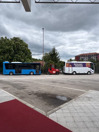 The image shows a parking area adjacent to a sidewalk in Nikšić, Montenegro. The area includes a blue bus and a white bus with purple writing which is parked next to a red vehicle. There is overcast weather in the area.