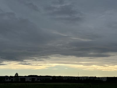 A silhouetted landscape of suburban homes and trees is visible against a dramatic sky filled with dark, heavy clouds. The clouds are layered and textured, with streaks of lighter gray and white suggesting movement and depth. The setting appears to be at dusk or during a storm, as indicated by the muted, somber lighting and the overcast sky. Below the sky, the horizon is lined with dark shapes of houses and a dense band of trees, all rendered in silhouette. A few structures stand out with lighter facades, but their details are obscured by the low light. In the foreground, a dark field or lawn is visible, with a few blurred plants or blades of grass suggesting the ground. The overall mood is somewhat melancholic or dramatic due to the imposing sky and the silhouetted environment. The location is identified as Harkujärve, Estonia, suggesting a Northern European setting. There is no visible text in the image.