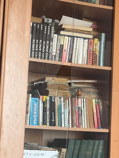 This image shows a bookshelf filled with various books. The bookshelf is made of wood and has glass doors. The books are arranged in rows and are of different sizes and colors. Some of the books have titles in Russian. The top shelf has several stacks of papers and a small object that appears to be a camera. The middle shelf is packed with books, some of which are leaning to the side. The bottom shelf also contains books. The overall impression is of a well-used and perhaps somewhat cluttered personal library. The image does not provide any information about the location in the city of Gari, Russia, or any specific details about people, actions, or interactions. It is a static scene of a bookshelf.