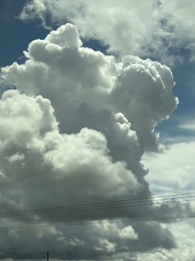 The image displays a sky scene over Kiryat Gat, Israel. Dominating the upper and central portions are large, bright white cumulus clouds, exhibiting significant vertical development and distinct, rounded contours. Interspersed among these and visible in the background are patches of clear, light blue sky. Towards the lower third of the image, the clouds appear darker and grayer, suggesting a greater depth or less direct sunlight penetration. Horizontally spanning across the lower half of the frame are five parallel, dark-colored power lines. A section of a dark utility pole is visible at the bottom center, extending upwards from off-frame, supporting the lowest of these wires. No individuals or specific ground features are discernible beyond the utility infrastructure.