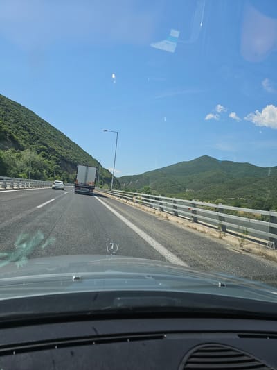 The image presents a forward-facing view from inside a vehicle traveling on a highway through a mountainous region. The sky is bright blue with a few scattered white clouds, indicating clear, sunny daytime weather.
On the right side of the frame, parts of the vehicle's interior are visible, including a dark dashboard element, an air vent, and the hood featuring a prominent silver Mercedes-Benz logo. Reflections on the windshield suggest interior details and potentially a document or sticker, though any text is illegible.
The highway is made of asphalt with clear white lane markings, including a solid line along the left edge and a dashed line separating the right lane. A large white and grey semi-trailer truck is visible ahead in the right lane, with its rear facing the camera, followed by a smaller white car.
To the left of the road, a substantial metal crash barrier runs alongside, beyond which steep, green mountains covered in dense foliage rise dramatically. Several streetlights are spaced along the left side of the highway, positioned between the road and the mountains. The scene depicts travel through a scenic, elevated landscape, consistent with the given location context of Promachonas Municipal Unit, Greece.