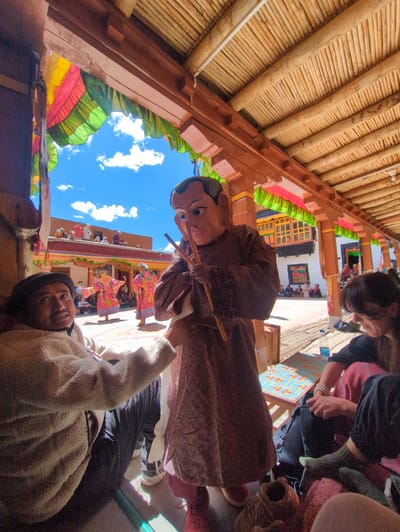 In the city of Pibiting, India, a lively outdoor gathering is underway, possibly at a monastery, as indicated by the architectural style and the presence of people in traditional attire. The scene is framed by a wooden roof structure supported by pillars, adorned with colorful fabric banners in red, yellow, and green. Below, a person in a beige, fuzzy jacket sits facing forward, looking up with a slight smile. In front of them, a figure wearing a large, mask-like headpiece with wide eyes and a pointed nose stands holding two wooden sticks. This figure is clad in a brown robe. In the background, two individuals in bright, ornate pink and yellow costumes are performing a dance, their movements captured mid-motion. A crowd of spectators sits or stands around the courtyard, observing the festivities. The sky above is a brilliant blue, dotted with white clouds, suggesting a clear, sunny day. The ground appears to be paved.
