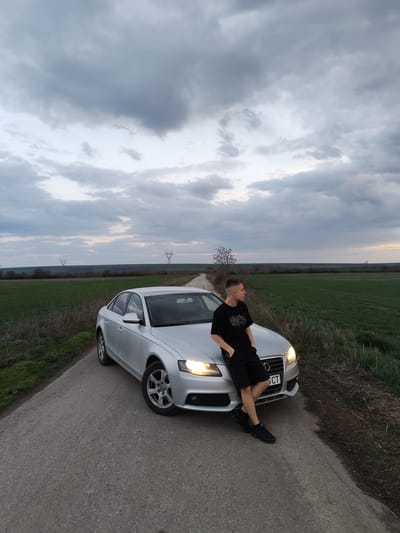 The image portrays a rural road in Gorni Tsibar, Bulgaria, under an overcast sky with predominantly gray clouds. A silver Audi A4 sedan is parked diagonally on a narrow, asphalt road, facing toward the viewer's right. Its headlights are illuminated. A young adult male is leaning against the front hood of the vehicle, facing right and looking away from the camera. He wears a black short-sleeved t-shirt with a white graphic, black shorts, and black athletic shoes. His right hand is in his pocket, and his left arm rests on the car's hood. The car's front license plate is partially visible, displaying "5 CT". Green agricultural fields border both sides of the road. In the mid-distance, a bare tree stands near a slight curve in the road. Further in the background, two high-voltage electricity pylons are visible against the horizon.