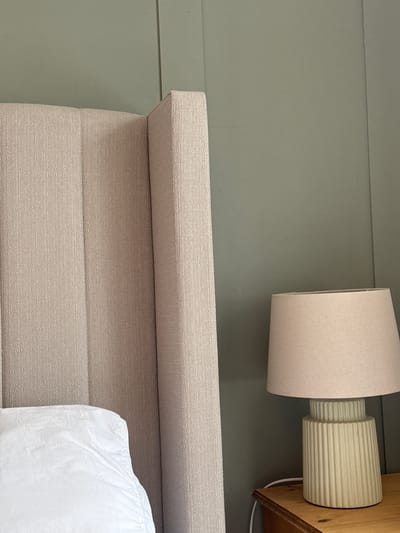 The image depicts a close-up of a bedroom interior. A beige fabric headboard with horizontal stitching is prominently featured in the lower half of the frame. A portion of a white pillow rests against the right side of the headboard.
Above the headboard, mounted on a pale green wall with horizontal paneling, is a wall lamp. The lamp has a cylindrical beige lampshade and a textured, fluted base that attaches to a wooden mounting.
The scene appears to be a detail shot of a modern or contemporary bedroom. No people are visible in the image. The precise location in Windermere, United Kingdom, cannot be determined from the visual information provided.