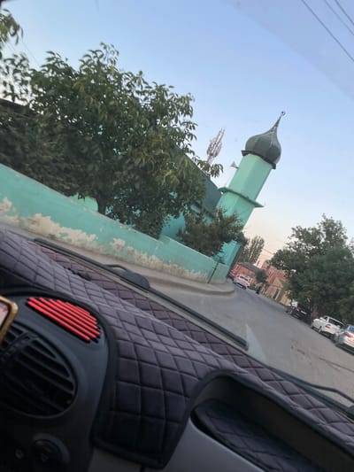 The image shows a street scene in Buynaksk, Russia, with a prominent turquoise-colored mosque with a green dome on the left side of the frame. A tall, slender minaret of the mosque extends into the clear blue sky. Trees with lush green foliage line the street, and the turquoise wall of the mosque, appearing somewhat weathered and peeling in places, runs along the left side. In the foreground, the interior of a vehicle is visible, including the dashboard with a quilted black cover and a red grille vent. Several cars are parked and driving on the asphalt road. The overall impression is a snapshot of urban life in Buynaksk, capturing architectural elements and the everyday flow of traffic.