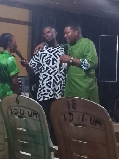Three people stand in a room. One woman in a green outfit holds a microphone. To her right, a man in a black and white patterned shirt looks at her. A second man in a green outfit has his arm around the man in the patterned shirt and looks toward him. In the foreground, two empty chairs have “AD.U.UM” written on them along with the numbers 6 and 18.