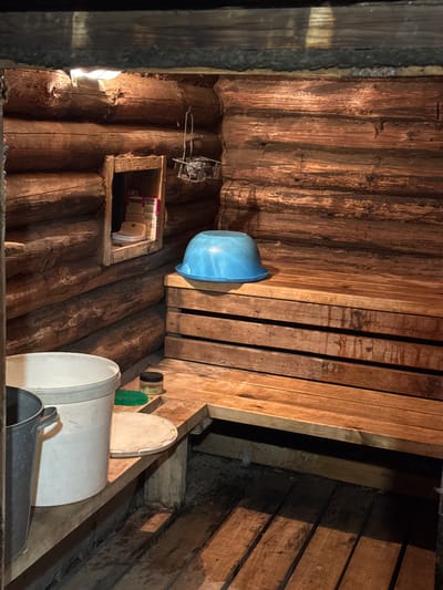 The image depicts the interior of a rustic wooden sauna, likely located in Maksimovo, Russia. The walls are constructed from rough-hewn logs. In the foreground, a large white bucket and a grey metal bucket are visible. To the right of the buckets, on a wooden shelf, is a small green scrubbing brush and a jar with a black lid. Behind these items, a light blue plastic basin rests on a wooden bench. The bench itself is made of several horizontal wooden planks. Further back and to the left, a small wooden shelf is built into the wall, containing what appears to be a white soap dish and a box labeled "TEMOC". Above this shelf, a metal basket containing stones hangs from the wall. A light fixture is visible on the ceiling logs, casting illumination on the scene. The wooden floorboards are visible in the lower portion of the image, appearing wet. The overall impression is of a functional, traditional sauna environment. There are no people present.