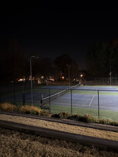 A nighttime image shows a tennis court illuminated by artificial lights. The court is enclosed by a dark fence. A row of tall ornamental grasses and shrubs separates the court from a dark retaining wall topped with dried grass. In the background, trees and houses are visible, dimly lit by street lamps and house lights. The left side of the image is dominated by darkness.