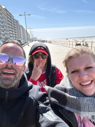 This bright, upright group selfie features three individuals enjoying a sunny day at a coastal location, likely Middelkerke, Belgium.
In the foreground on the left, a bearded man with light blue mirrored sunglasses smiles broadly, revealing his teeth. He is wearing a dark hooded jacket. Next to him, a woman with dark hair wears dark sunglasses, a red baseball cap, and a black hooded jacket over a red and white striped top. She poses playfully with a pout and makes a peace sign with her right hand, adorned with bright pink nail polish. To her right, a blonde woman smiles widely, wearing a grey patterned scarf and a dark jacket, possibly over a pink top.
The background reveals a vibrant outdoor scene. Behind the individuals is a wide, paved promenade, with several parked bicycles visible on the far right. Beyond the promenade lies an expansive sandy beach, stretching towards the sea under a clear blue sky. Faint outlines of beach chairs or cabins are visible in the distance on the beach. To the left of the subjects, a row of tall, modern multi-story buildings with numerous windows lines a street, with streetlights and parked cars visible in front of them.
The bright conditions indicate it is daytime, likely midday or early afternoon, with clear, sunny weather. The subjects appear happy and relaxed, enjoying their outing. No discernible text is visible on clothing or in the background.