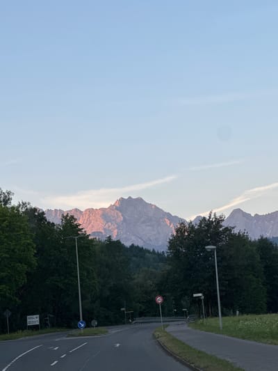 The image captures a scenic view of mountains bathed in the warm glow of sunset or sunrise, suggesting either early morning or late evening. The sky is a clear, pale blue, with wispy clouds stretching across the horizon. In the foreground, a dense forest of green trees lines the side of a paved road. The road curves gently to the right, with a grassy verge and a low fence bordering it.
Several objects are visible along the roadside. A tall lamppost stands prominently, and further down, there are road signs. One sign is circular with a red border and the number "60" in black, indicating a speed limit. Another sign is blue with a white arrow pointing upwards, signifying a direction. A smaller sign with text is also visible near the fence.
The overall scene evokes a sense of tranquility and natural beauty. The rugged mountain peaks contrast with the lush greenery of the forest and the man-made road. The warm light on the mountains adds a dramatic and serene quality to the landscape. The presence of road signs suggests this is a public road, likely in a rural or semi-rural area, given the natural setting. The location context of Farchant, Germany, places this scene within the Bavarian Alps region.
