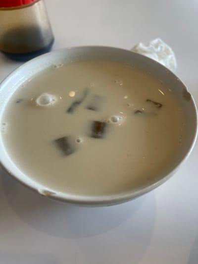 A white bowl filled with a creamy, beige liquid and dark, cubed pieces is centered in the frame, resting on a white surface. The liquid has small bubbles on its surface, and the cubes appear to be floating within it. In the upper left corner, a glass bottle with a dark liquid and a red cap is partially visible. To the upper right of the bowl, a crumpled white napkin is present. The scene is well-lit, suggesting an indoor setting. The white surface on which the bowl sits has a shadow cast from the bowl, indicating a light source from the upper right. There are no people visible in the image.