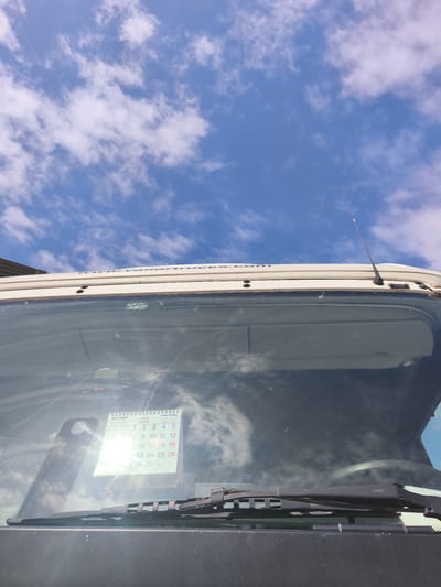 This media file captures a bright outdoor scene looking upwards from the perspective of a vehicle's interior or immediately in front of it, against a clear blue sky with scattered white clouds. The primary subjects are the front of a vehicle, partially obscured by its windshield, and the sky above.
The foreground is dominated by the vehicle's windshield, which reflects the sky and clouds. Below the windshield, the top of the dashboard or hood is visible, along with black windshield wipers resting horizontally. Above the windshield, the curved white roof of the vehicle is seen, featuring what appears to be part of a logo or text, partially readable as "www. vatterfrucks". An antenna extends upwards from the top right side of the vehicle's roof. Through the windshield, sun visors are discernible, and a small sticker with "SPAIN" and other text is visible near the top. Another fragmented text, "A-85L-20 KWH", is also seen, likely from a reflection or another sticker.
Prominently displayed on the inner side of the windshield is a small white spiral-bound calendar. The calendar clearly indicates "Julio 2026" (July 2026) in Spanish. The days of the month are laid out, showing the 1st through the 5th, then the 7th through the 12th, followed by the 15th through 19th, 23rd through 26th, and finally 29th through 31st. The 1st of July appears to fall on a Wednesday.
The overall impression is one of a sunny day in Nàquera / Náquera, Spain, with the vehicle parked outdoors. The bright lighting suggests it is daytime.