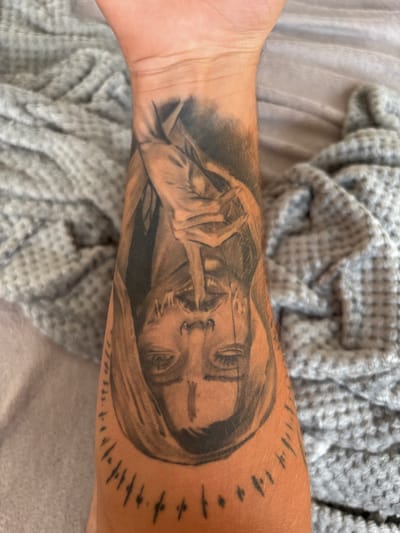 The image shows a tattoo on a person's forearm. The tattoo depicts a person's face with a hand covering their mouth, rendered in a black and grey style. The tattoo is oriented upside down relative to the viewer. The forearm is resting on a grey, textured blanket. There are no observable elements that indicate the location in Stadtroda, Germany.