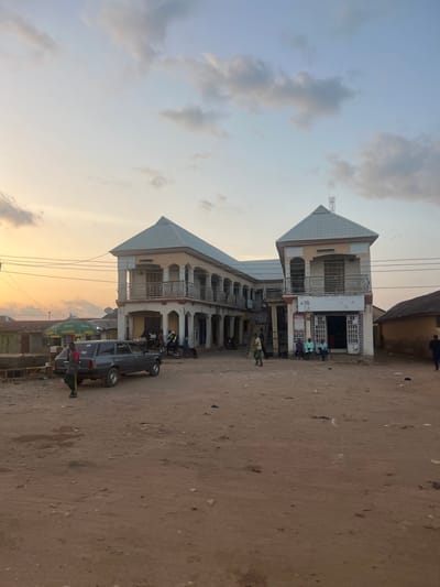 This image depicts a late afternoon or early evening scene in Rinjim Gani, Nigeria. The sky transitions from a warm orange-yellow near the horizon to a pale blue overhead, with scattered light grey clouds. The ground consists of uneven reddish-brown dirt, showing signs of vehicle passage and scattered small debris.
A two-story building dominates the central-right portion of the frame. It features a light-colored facade with white trim, a light grey pyramidal roof, and a small antenna mast at its apex. The ground floor displays multiple arched entryways, and the upper floor has an open balcony with metal railings. A shop on the ground floor, beneath the right side of the balcony, has open double doors and a sign reading "BAZAN AUTO COSMETICS." The number "16" is visible on the building's facade on the left side of the ground floor.
To the left of the main building, a dark grey Peugeot 504 station wagon is parked on the dirt, facing right. A person in a dark striped shirt and green pants stands next to the driver's side of the car, facing away. Further left, behind the car, are smaller, single-story structures, some with dark roofs, and two prominent green market umbrellas. Overhead power lines are visible spanning across the left and central parts of the image.
Several individuals are present: three men are seated on chairs outside the "BAZAN AUTO COSMETICS" shop; a person in light attire stands near the center of the main building; and another person in dark clothing walks towards the right side of the frame. Two individuals are also visible on a motorcycle parked near the left side of the main building. To the right of the main building, a single-story structure with a dark, sloped roof is partially visible.