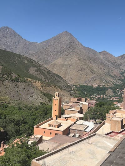 A wide, high-angle view captures a mountain village in the Asni region of Morocco under a clear blue sky. The foreground features a cluster of earthen-toned buildings with predominantly flat roofs. A prominent, tall, rectangular minaret, colored orange-brown, stands centrally, displaying decorative arched openings near its peak. Adjacent to it is a building with a lighter-colored flat roof and a small, square, raised structure with multiple arched windows. Various other buildings are visible, some with exposed rebar and unfinished concrete slabs on their roofs, indicating construction or renovation. The midground presents a deep, verdant valley filled with dense tree cover, within which smaller clusters of buildings are visible. A winding road ascends the slopes in the distance. The background is dominated by large, rocky mountains, primarily gray and brown, with sparse green vegetation. The peak on the far left shows small patches of light-colored rock or snow. The sky is a vast, cloudless blue, with a faint crescent moon visible in the upper right quadrant. The scene is brightly lit by direct sunlight, casting defined shadows. No people, animals, or vehicles are present.