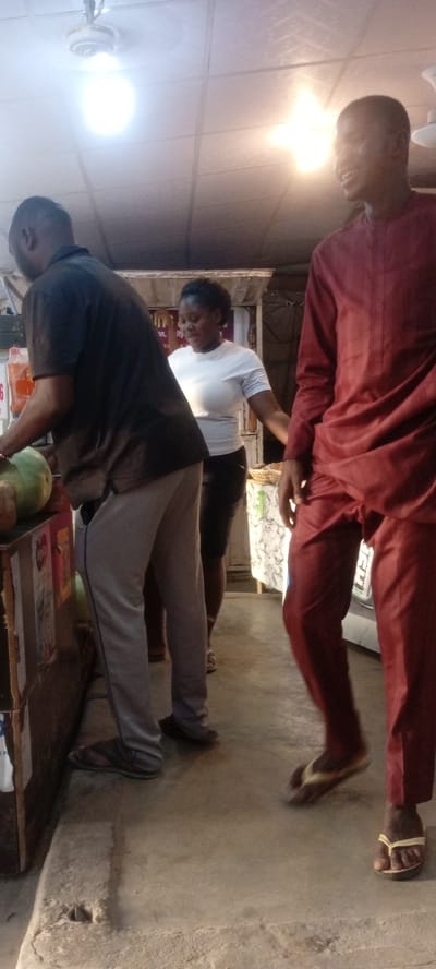 In a brightly lit indoor space in Rijiyar Zaki, Nigeria, three individuals are present. On the left, a man dressed in a black shirt and grey pants stands at a counter where fruits, including watermelons, are displayed. A woman in a white shirt and black shorts stands in the middle. To the right, a man in a red, formal outfit, coupled with flip-flops, smiles as he walks. Overhead, two ceiling lights and a fan illuminate the scene.