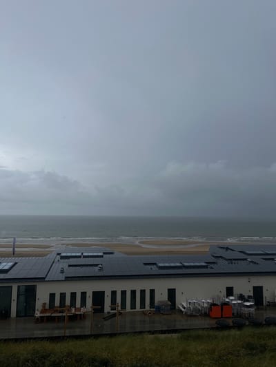 The image depicts a coastal scene in Overveen, Netherlands, under a heavily overcast, dark grey sky. The upper two-thirds of the frame are dominated by dense, low-hanging clouds. In the mid-ground, a wide, grey-brown sandy beach with visible wave patterns from receding water stretches between the ocean and a building. The North Sea appears grey and choppy, with a faint horizon line.
In the foreground, a dark green, unkempt grassy slope partially obscures the base of a modern, single-story building with white exterior walls. The building features a dark, multi-faceted roof extensively covered with numerous black rectangular solar panels and several integrated skylights. The building's facade facing the viewer includes multiple tall, narrow dark-framed windows or doors, along with a larger glass door on the left side.
An outdoor patio or terrace, appearing wet and reflective, extends from the building. On the left side of this patio, a wooden outdoor dining table with benches is visible, surrounded by several vertical wooden posts. Miscellaneous items, including what appears to be building materials or furniture components, are present. On the right side of the patio, various supplies and equipment are stored: stacks of metal trolleys or racks holding white containers, two bright orange bins, and several large, dark, circular objects resting on the ground. No individuals are visible anywhere in the scene.