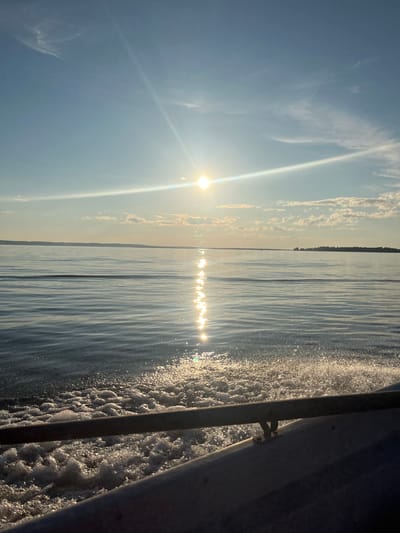 This image depicts a serene view of the wide, calm waters of Lake Ladoga near the village of Siukterka, Russia, bathed in the warm glow of the setting sun. The sun, a bright orb in the sky, casts a shimmering reflection across the water's surface, creating a dazzling path of light. Gentle ripples disturb the water, suggesting a slow-moving vessel, possibly a boat or a ferry, as evidenced by the foamy wake visible in the foreground. A metallic railing, likely part of the boat's structure, runs horizontally across the bottom of the frame, adding a sense of perspective and hinting at the viewer's vantage point. In the distance, a low, tree-lined horizon marks the distant shore. The sky is a pale blue, transitioning to a warmer hue near the horizon, with a few wispy clouds adding texture to the scene. The overall impression is one of tranquility and natural beauty, characteristic of a peaceful day on the lake.