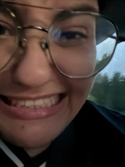 A close-up, slightly blurry, selfie-style photo shows a person's face from the chest up, with a smiling expression. The person is wearing large, round glasses. The background is out of focus and appears to be a natural landscape with trees and a cloudy sky, suggesting an outdoor setting. The exact location of Rygge, Norway is not discernible from the visual elements within the image.