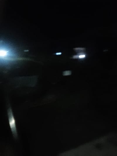 The scene depicts an indistinct, extremely low-light outdoor environment, identified as Njoro ward, Kenya, during nighttime. The image exhibits significant motion blur and digital noise throughout. Primary visible elements are various light sources against an almost entirely black background. A prominent, diffuse white light source is situated on the far left of the frame, illuminating a small, undiscernible area. Towards the mid-right, two distinct, elongated, horizontal light blue/white streaks are visible, alongside another fainter white streak further right. A subtle, rectangular, dim light appears in the lower-mid ground. A narrow, vertical streak of white light runs along the lower-left edge of the frame. No identifiable persons, specific objects, vehicles, or discernible actions or interactions are present within the captured scene.