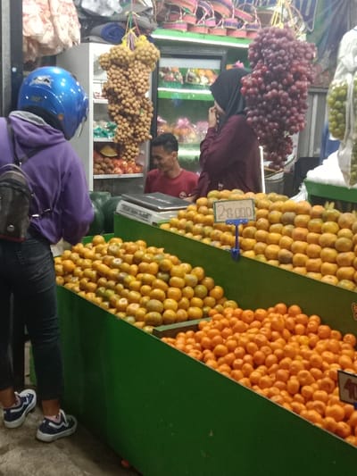 The image depicts an active fruit stall in a market located in Sekopek Wetan, Indonesia. In the foreground, a person is seen from the back, wearing a purple hooded sweatshirt, black jeans, and blue and white sneakers, with a blue motorcycle helmet on their head and a dark backpack. They are positioned in front of multiple green, slanted bins filled with piles of oranges or mandarins. A blue price tag displaying "28.000" is visible within one of the upper fruit bins. Behind the fruit display, a male stall worker in a red t-shirt is visible, smiling, standing behind a counter that holds a silver weighing scale. To his right, a female customer, partially obscured, wears a dark hijab and a maroon long-sleeved garment, looking towards the fruit. Hanging from above are large bunches of light brown, round fruit (likely longan) and dark red grapes, along with bags of green fruit. In the background, a refrigerated display case contains various packaged items, and woven baskets are hanging overhead. Several green watermelons are visible on the counter behind the foreground customer.