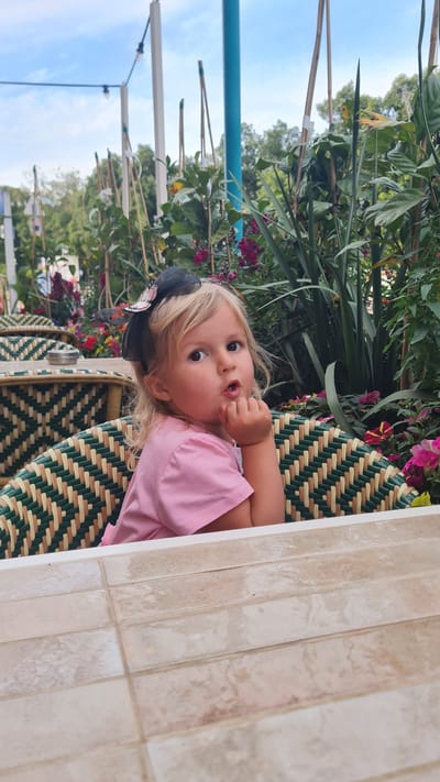 A young child with blonde hair and a black bow is sitting in a wicker chair at a table with tile-like surfaces. The child wears a pink shirt and is holding a hand to their mouth. Behind the chair are various green plants and pink flowers. In the background, there is a glimpse of a blue sky, green trees, and urban elements like poles and wires, suggesting the location may be in a city, such as Kiryat Gat, Israel.