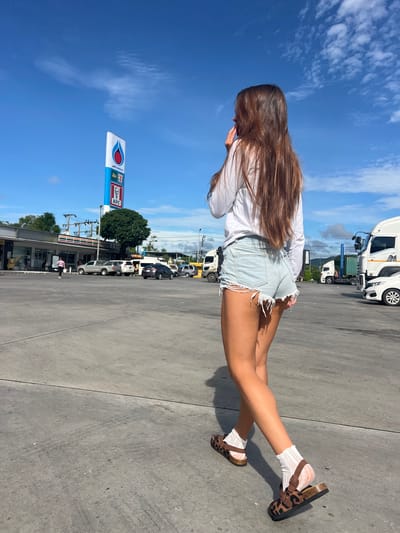 In the city of Ban Ko Pru, Thailand, a woman with long brown hair, wearing a white long-sleeved shirt and distressed denim shorts, walks across a concrete lot under a bright blue sky with scattered clouds. She has on leopard print sandals with white socks. Behind her, a PTT Station sign with a PTT logo, a 7-Eleven logo, and a KFC logo is visible, along with various vehicles including cars, trucks, and a white car in the foreground. A row of shops is also present on the left side of the image.