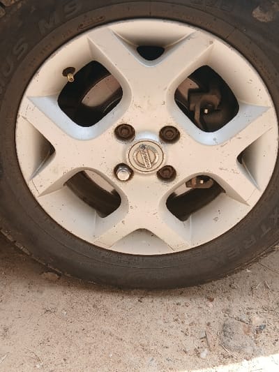 The image presents a close-up view of a vehicle's wheel and tire, situated in Mumbuni North ward, Kenya. The wheel consists of a five-spoke, silver-colored alloy rim, which appears soiled with dust and grime. A brass-colored valve stem, topped with a dark cap, protrudes from the rim. The central hub cap displays a tarnished Nissan logo. Surrounding the hub cap are five lug nuts: four are visibly corroded and brown, while one is silver. Through the openings in the rim's spokes, metallic brake components, specifically a rotor and caliper assembly, are partially visible. The tire sidewall prominently features the brand name "MAXTREK," along with additional markings including "PLUS MS" and "TUBELESS." The wheel is resting on a dry, light-brown ground surface composed of sand and dirt, intermingled with scattered small stones and minor debris. No individuals or specific actions are depicted within the frame.